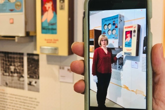 Ein Smartphone wird in der Ausstellung in der Hand gehalten und zeigt ein Video einer Museumsmitarbeiterin, welche den fahinter hängenden Automaten erklärt.