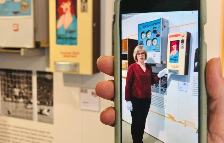 Ein Smartphone wird in der Ausstellung in der Hand gehalten und zeigt ein Video einer Museumsmitarbeiterin, welche den fahinter hängenden Automaten erklärt.