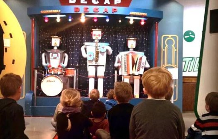 Eine Gruppe von Erstklässlern sitzt mit dem Rücken zur Kamera auf einem roten Teppich und blivkt auf eine Band von drei Robotern, welche Musikinstrumente spielen