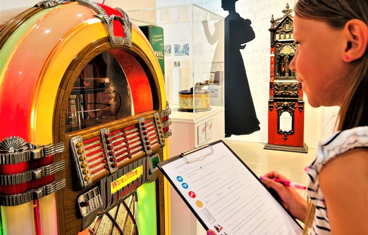 Ein Mädchen im Profil blickt auf die 40er-Jahre Jukebox Wurlitzer 1015 und hält einen Quizbogen und Stift in den Händen.